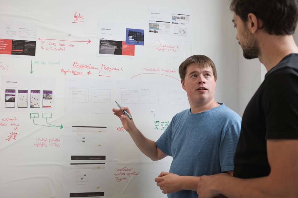 Two men stand in front of a white board and discuss a digital transformation project