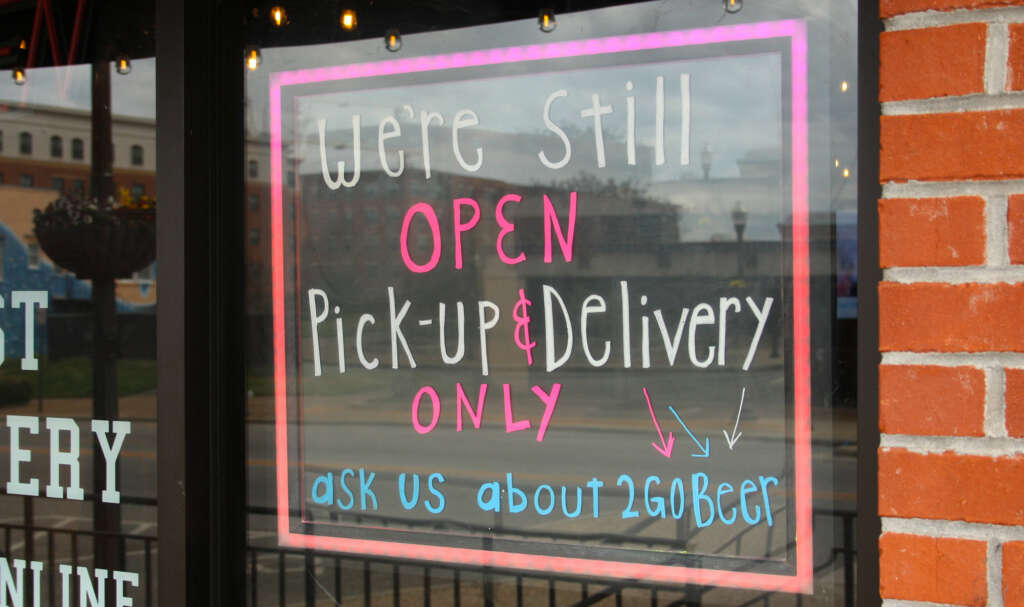 A sign in the window of a bar says it is still open of pick up and delivery