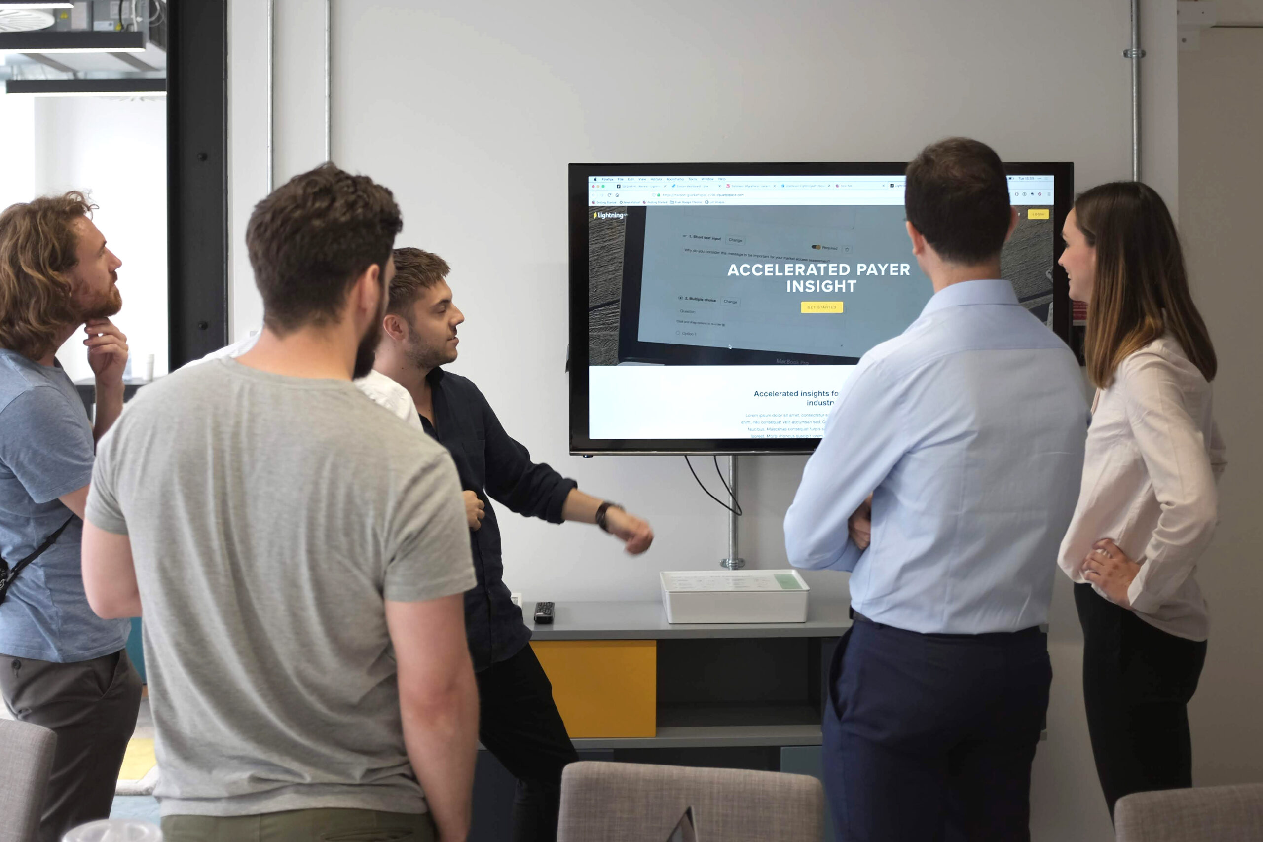 A group of people stand around a screen and discuss the design of a website