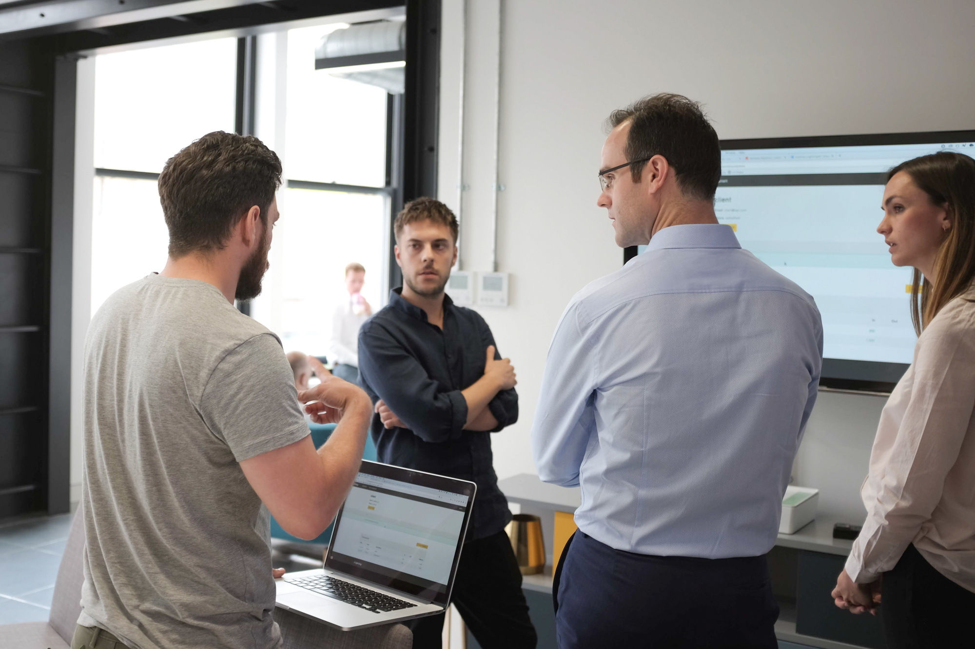 A group of people are having a stand up meeting around a screen