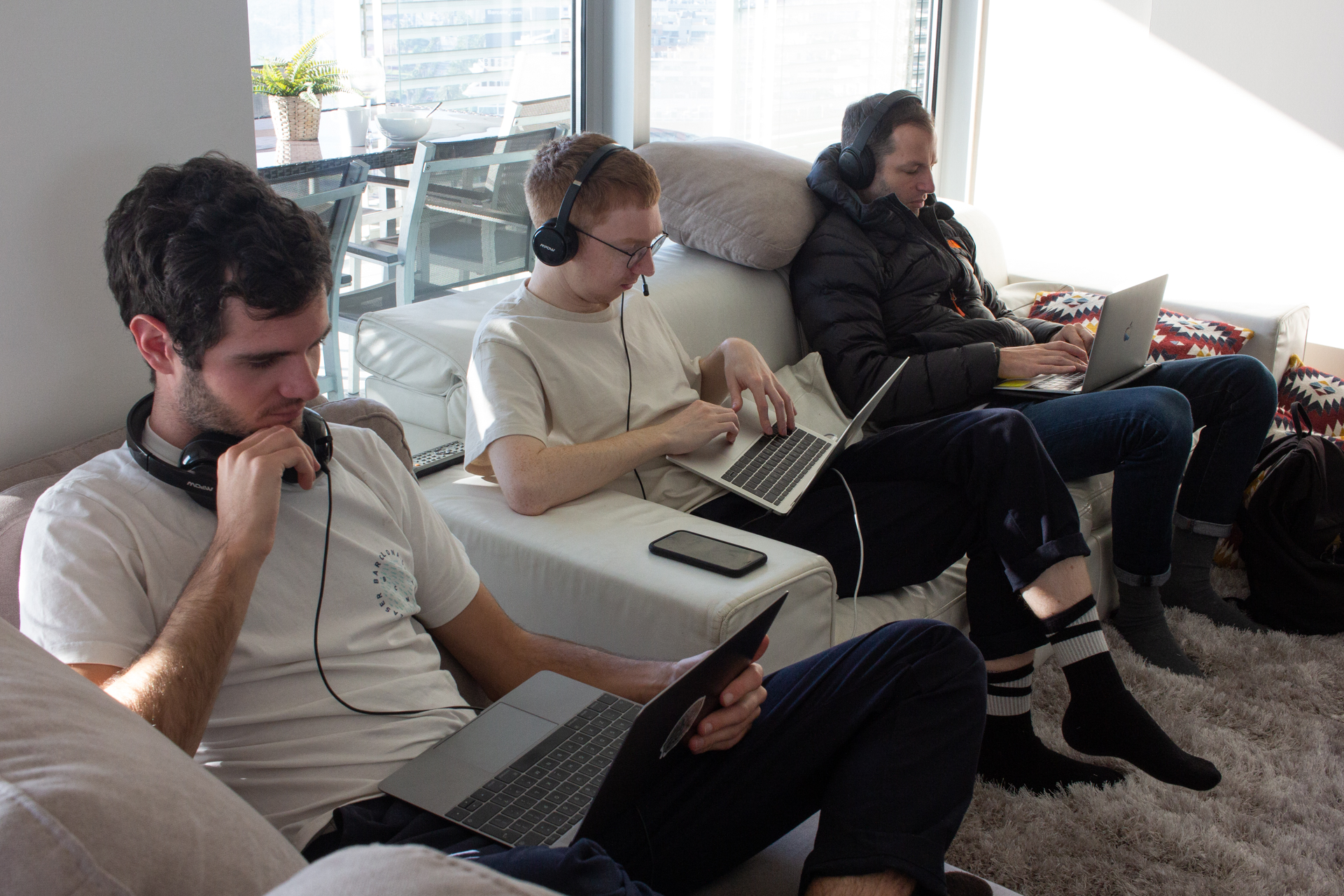 A team of coders work on laptops on a sofa in an apartment.